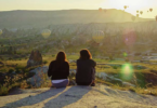 two people sitting on a hill looking at hot air balloons