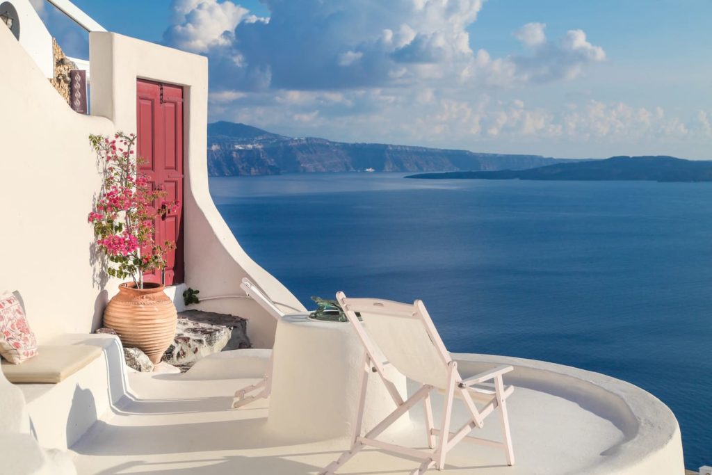 a white building with a red door and a pot with flowers on a balcony overlooking the ocean