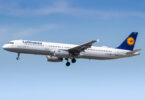 An airplane in flight against a clear blue sky. The aircraft is white with a blue tail fin featuring a yellow logo. The landing gear is extended, and the engines are visible under the wings. The word "Lufthansa" is written on the side of the plane.