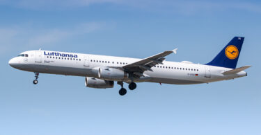An airplane in flight against a clear blue sky. The aircraft is white with a blue tail fin featuring a yellow logo. The landing gear is extended, and the engines are visible under the wings. The word "Lufthansa" is written on the side of the plane.