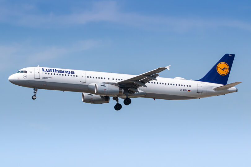 An airplane in flight against a clear blue sky. The aircraft is white with a blue tail fin featuring a yellow logo. The landing gear is extended, and the engines are visible under the wings. The word "Lufthansa" is written on the side of the plane.