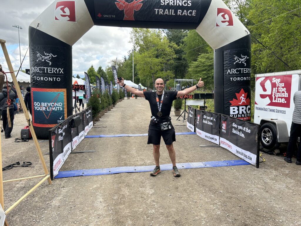 A man is standing at the finish line of a trail race, smiling and giving a thumbs-up. He is wearing a race bib and a medal around his neck. The finish line arch has logos and text, including "Arc'teryx Toronto" and "See you at the finish line." There are banners and a timing clock in the background, along with trees and a cloudy sky.