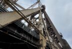 The image shows a close-up view of a large steel bridge structure, featuring intricate metal beams and cables. The perspective is from below, looking up at the bridge, highlighting its architectural details against a cloudy sky. There are fences and railings visible at the bottom of the image.