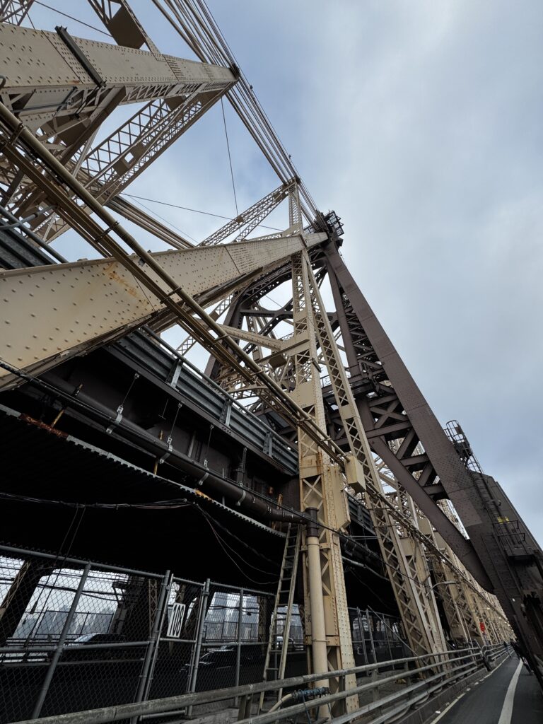 The image shows a close-up view of a large steel bridge structure, featuring intricate metal beams and cables. The perspective is from below, looking up at the bridge, highlighting its architectural details against a cloudy sky. There are fences and railings visible at the bottom of the image.