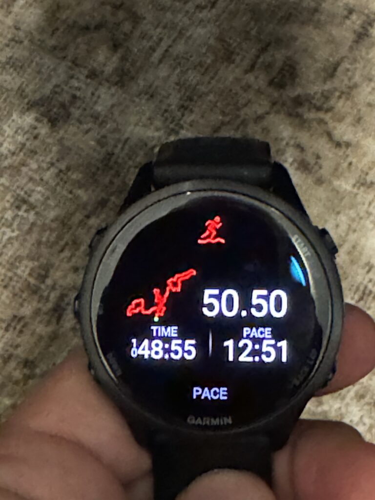 A person is holding a Garmin smartwatch displaying workout data. The screen shows a red path map, a distance of 50.50, a time of 48:55, and a pace of 12:51. The background is a textured surface.