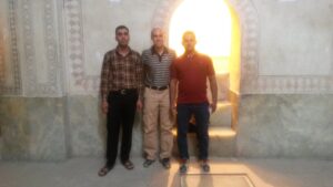 Three men are standing side by side in front of an ornate wall with decorative patterns. There is a bright, arched doorway behind them, illuminating the scene. The men are dressed in casual attire, with one wearing a plaid shirt, another in a striped shirt, and the third in a solid-colored shirt. The setting appears to be indoors, possibly in a historical or cultural building.