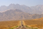 A long, straight road stretches into the distance, leading towards a range of rugged, brown mountains under a clear blue sky. A single vehicle is visible on the road, and utility poles line the right side. The landscape is arid and barren, with sparse vegetation.