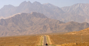A long, straight road stretches into the distance, leading towards a range of rugged, brown mountains under a clear blue sky. A single vehicle is visible on the road, and utility poles line the right side. The landscape is arid and barren, with sparse vegetation.