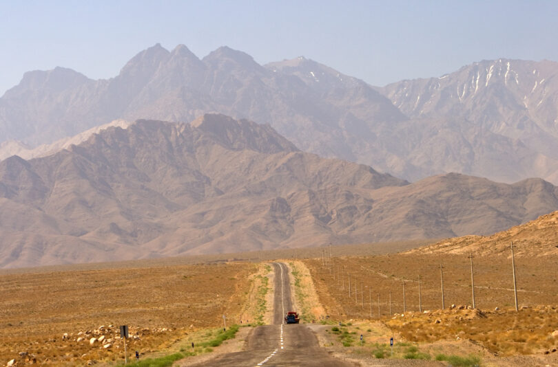 A long, straight road stretches into the distance, leading towards a range of rugged, brown mountains under a clear blue sky. A single vehicle is visible on the road, and utility poles line the right side. The landscape is arid and barren, with sparse vegetation.
