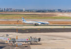 A commercial airplane with blue and white livery is taxiing on a runway at an airport. The tail of the plane features a blue Star of David. In the foreground, there is an airport terminal with a jet bridge labeled "C9" and signs in Hebrew. The background shows a cityscape with several buildings and a clear blue sky.