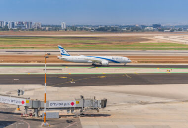 A commercial airplane with blue and white livery is taxiing on a runway at an airport. The tail of the plane features a blue Star of David. In the foreground, there is an airport terminal with a jet bridge labeled "C9" and signs in Hebrew. The background shows a cityscape with several buildings and a clear blue sky.