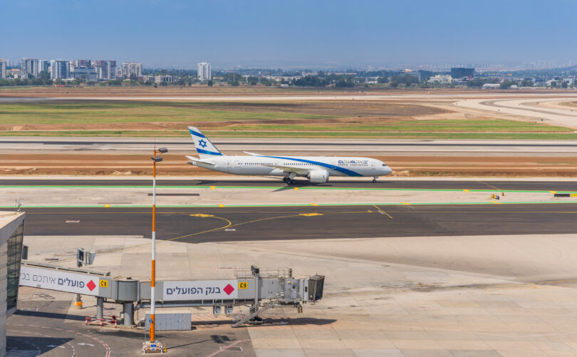 A commercial airplane with blue and white livery is taxiing on a runway at an airport. The tail of the plane features a blue Star of David. In the foreground, there is an airport terminal with a jet bridge labeled "C9" and signs in Hebrew. The background shows a cityscape with several buildings and a clear blue sky.