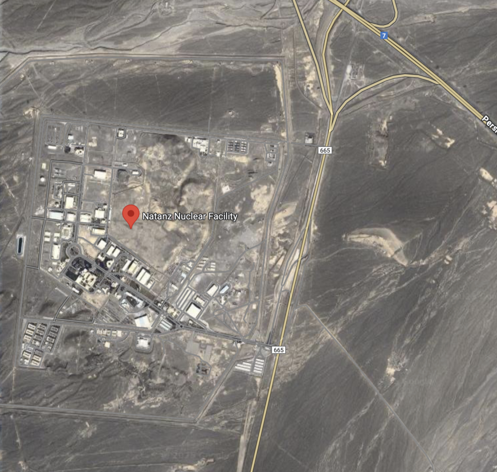 A satellite image showing a large industrial complex labeled "Natanz Nuclear Facility." The facility is situated in a desert area with surrounding roads and infrastructure. The landscape is mostly barren with some visible structures and buildings within the complex.