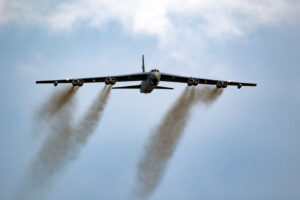 A large military aircraft is flying in the sky, viewed from below. It has a wide wingspan with multiple engines, emitting dark smoke trails. The sky is mostly clear with some clouds.