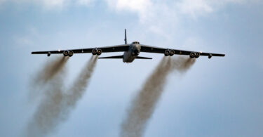 A large military aircraft is flying in the sky, viewed from below. It has a wide wingspan with multiple engines, emitting dark smoke trails. The sky is mostly clear with some clouds.