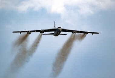 A large military aircraft is flying in the sky, viewed from below. It has a wide wingspan with multiple engines, emitting dark smoke trails. The sky is mostly clear with some clouds.