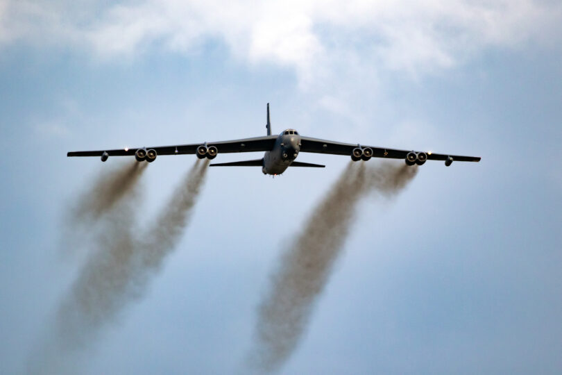A large military aircraft is flying in the sky, viewed from below. It has a wide wingspan with multiple engines, emitting dark smoke trails. The sky is mostly clear with some clouds.