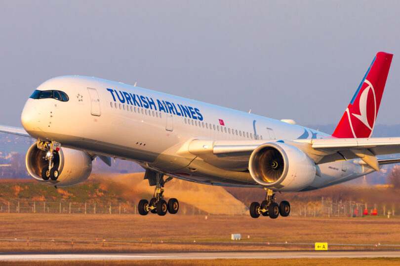 A Turkish Airlines airplane is landing on a runway. The aircraft is white with the airline's logo and name visible on the fuselage. The engines and landing gear are clearly visible, and the background shows a grassy area and a clear sky.