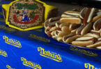 A championship belt is displayed on a table covered with a blue tablecloth featuring the Nathan's logo. Next to the belt, there is a large stack of hot dogs in buns. The background shows a brick wall with more Nathan's signage.
