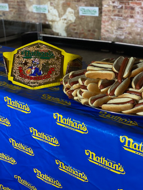 A championship belt is displayed on a table covered with a blue tablecloth featuring the Nathan's logo. Next to the belt, there is a large stack of hot dogs in buns. The background shows a brick wall with more Nathan's signage.