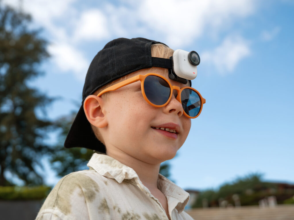 A young child is wearing a black cap with a small camera attached to it. The child is also wearing orange sunglasses and a light-colored shirt. The background features a clear blue sky and some blurred greenery.