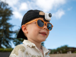 A young child is wearing a black cap with a small camera attached to it. The child is also wearing orange sunglasses and a light-colored shirt. The background features a clear blue sky and some blurred greenery.