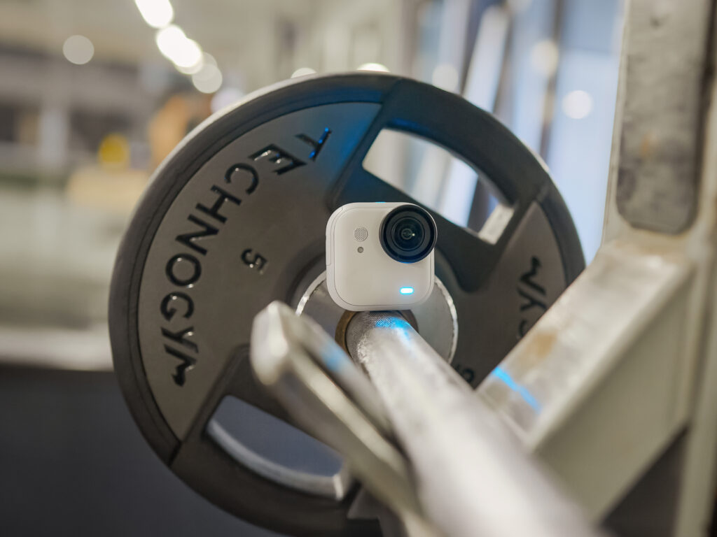 A small white action camera is mounted on a barbell in a gym setting. The barbell has a weight plate with the brand name "TECHNOGYM" visible. The background is blurred, suggesting a gym environment.