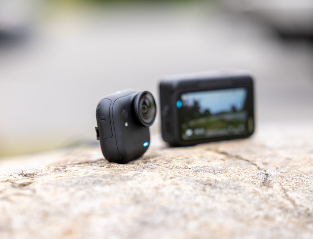 A small black action camera with a lens is placed on a rock surface. In the background, another camera with a screen displaying a blurred outdoor scene is visible. The image is focused on the first camera, with a soft, out-of-focus background.
