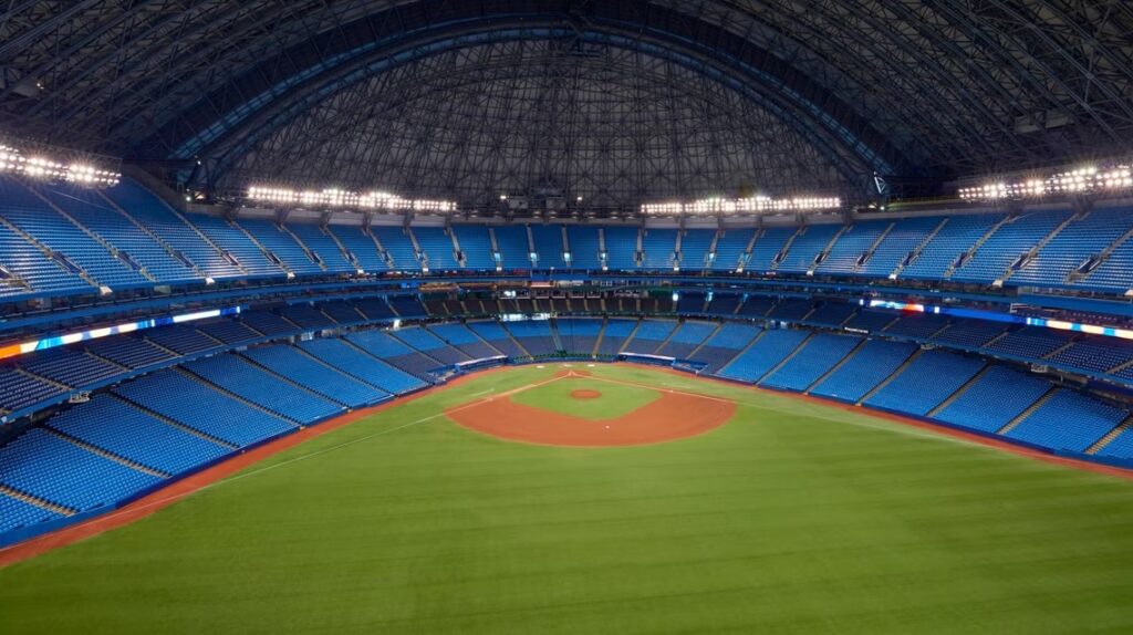 The image shows an empty indoor baseball stadium with a large, domed roof. The seating areas are filled with blue seats, and the field is well-maintained with green grass and a red clay infield. Bright stadium lights illuminate the space.