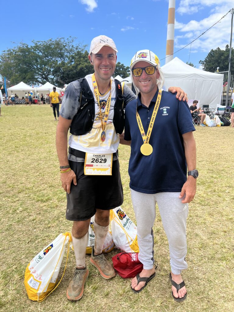 Two men are standing on grass, smiling at the camera. The man on the left is wearing a cap, a running vest, a race bib numbered 2629, and has a medal around his neck. The man on the right is wearing a cap, sunglasses, a dark shirt, light pants, and flip-flops, also with a medal. They are in a park-like setting with tents and people in the background.