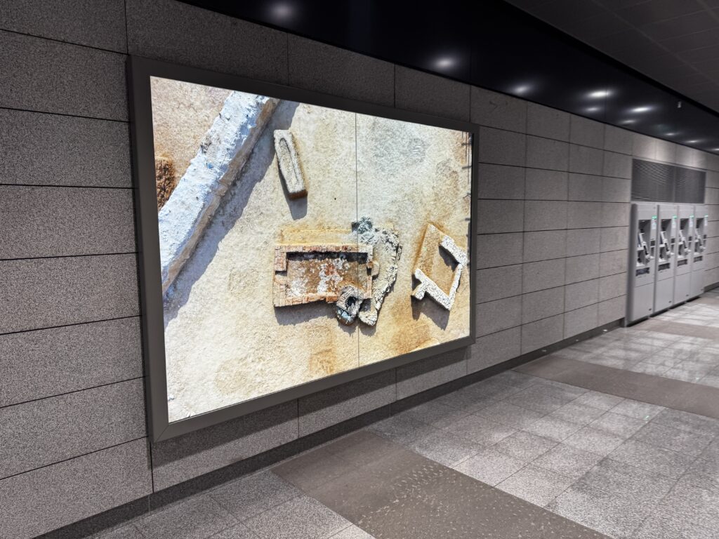 A large framed image is displayed on a tiled wall in a modern indoor setting, possibly a subway or train station. The image shows an aerial view of an archaeological site with various stone and brick structures partially buried in sand. The floor is made of gray tiles, and there are ticket machines or kiosks visible in the background.