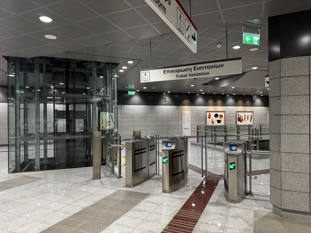 The image shows the interior of a modern subway station. There are ticket validation turnstiles with digital displays and green arrows indicating entry. Above the turnstiles, there is a sign in Greek and English that reads "Ticket Validation." To the left, there is a glass elevator shaft. The walls are lined with light gray tiles, and there are framed artworks displayed on the wall in the background. The ceiling is fitted with recessed lighting, and there are directional signs and an emergency exit sign visible.