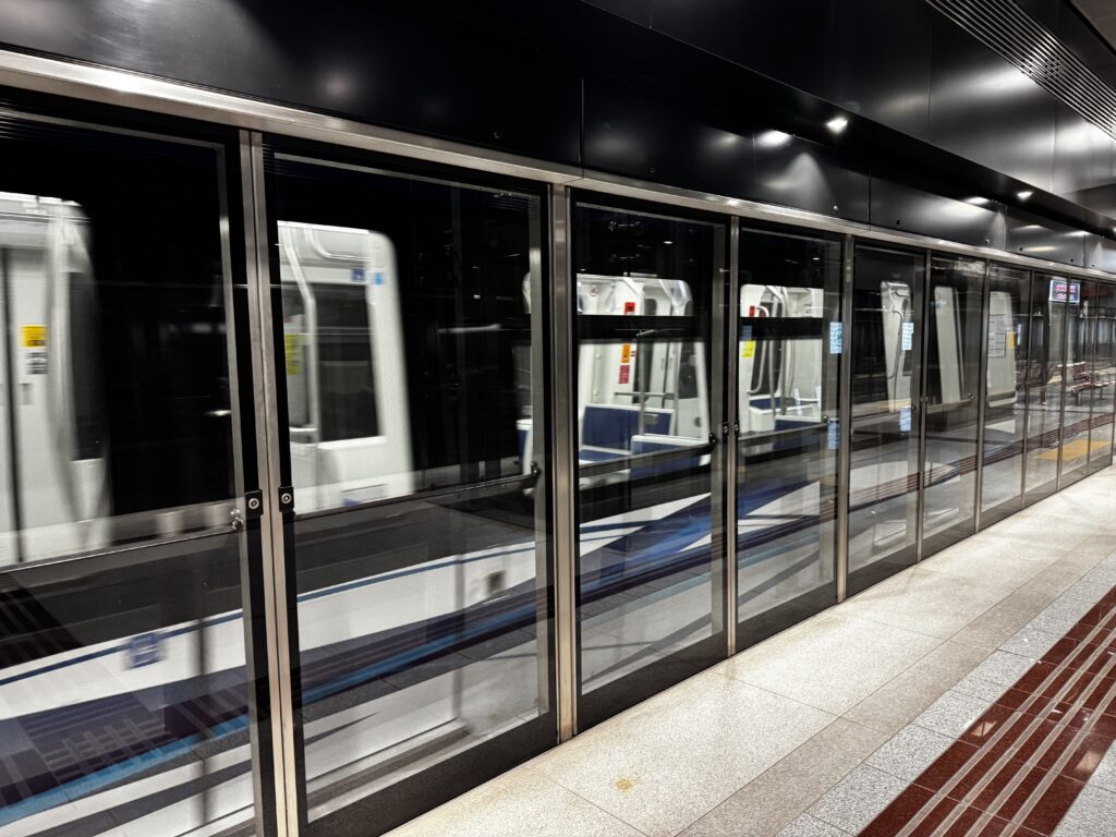 A subway train is moving past a platform with glass safety doors. The train is blurred, indicating motion, and the platform is empty with a clean, modern design. The lighting is bright, and the floor has a polished appearance.