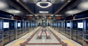 The image shows a modern, empty subway station platform. The platform is lined with glass doors and digital screens displaying a logo. There are red benches in the center, and the ceiling features circular light fixtures. The floor has tactile paving for accessibility, and an electronic sign displays information above. The station has a sleek, clean design with reflective surfaces.
