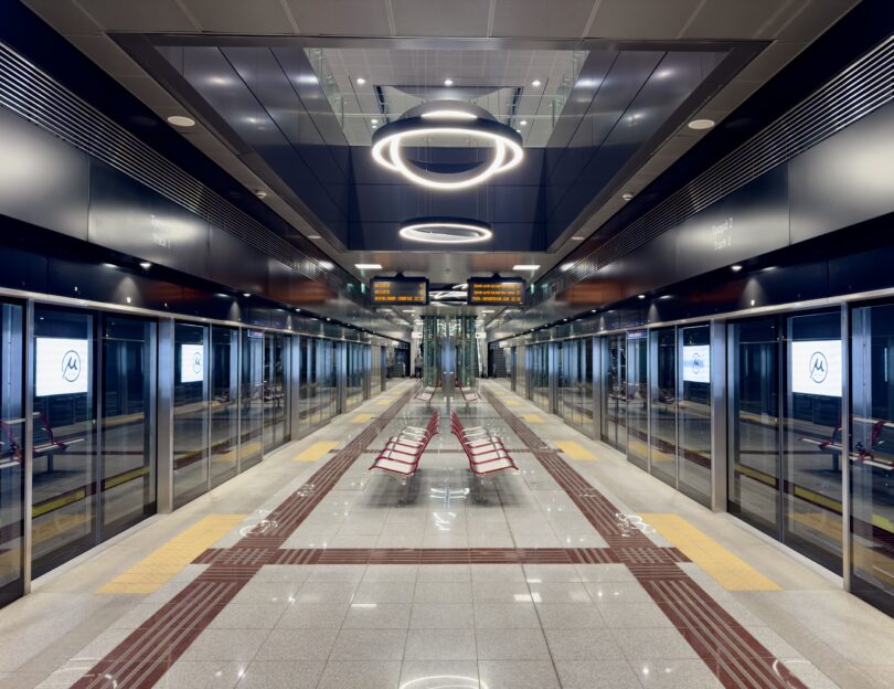 The image shows a modern, empty subway station platform. The platform is lined with glass doors and digital screens displaying a logo. There are red benches in the center, and the ceiling features circular light fixtures. The floor has tactile paving for accessibility, and an electronic sign displays information above. The station has a sleek, clean design with reflective surfaces.