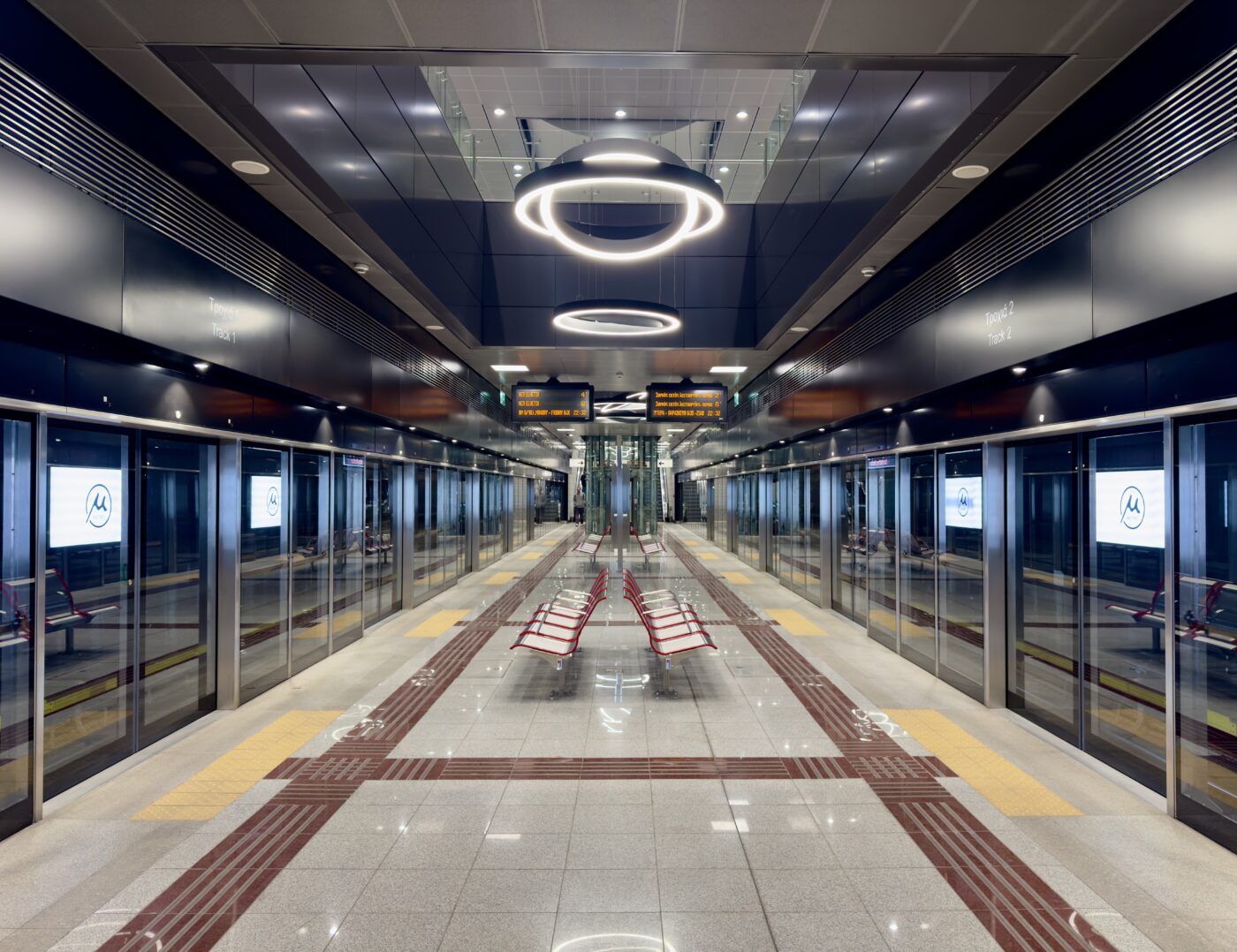 The image shows a modern, empty subway station platform. The platform is lined with glass doors and digital screens displaying a logo. There are red benches in the center, and the ceiling features circular light fixtures. The floor has tactile paving for accessibility, and an electronic sign displays information above. The station has a sleek, clean design with reflective surfaces.