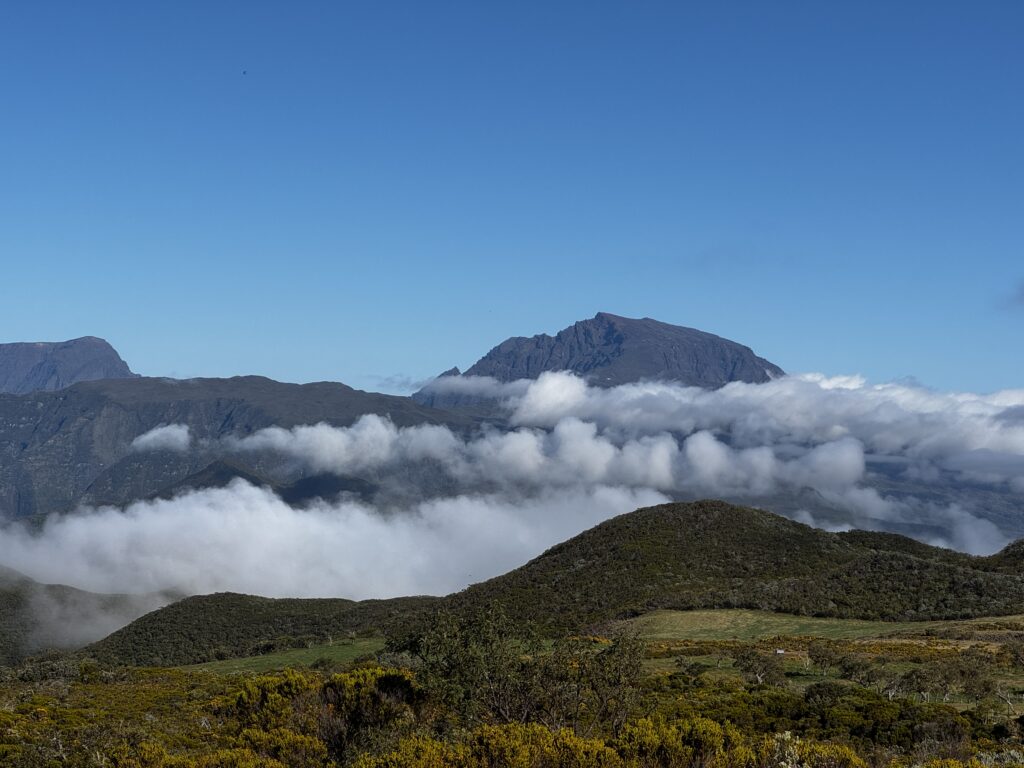 A scenic view of a mountainous landscape with lush green hills in the foreground. White clouds are drifting across the middle of the image, partially obscuring the distant mountains. The sky is clear and blue, adding to the serene atmosphere.