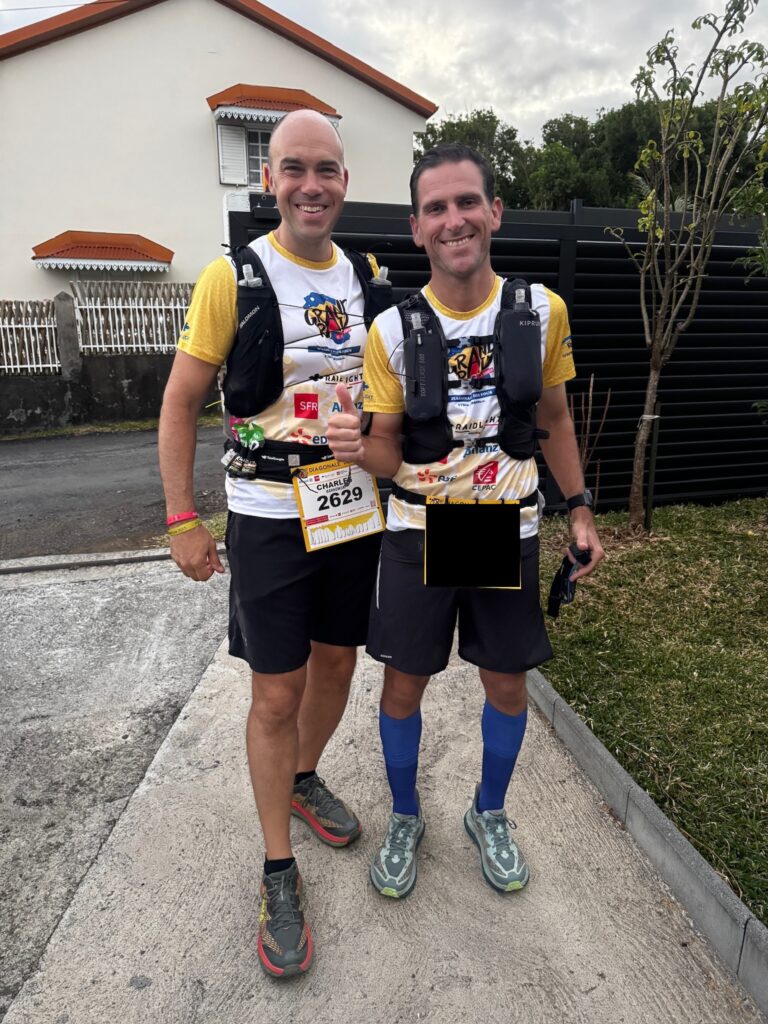 Two men are standing side by side, smiling at the camera. They are wearing matching athletic gear, including yellow and white shirts with various logos, black shorts, and running shoes. Both have hydration packs on their backs. One man is giving a thumbs-up. They are standing on a paved path with a house and some greenery in the background.