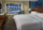 The image shows a hotel room with a large bed, a TV mounted on the wall, a desk with a chair, and a small armchair. The room has a large window with a view of a sports stadium, showing rows of blue seats and part of the field.