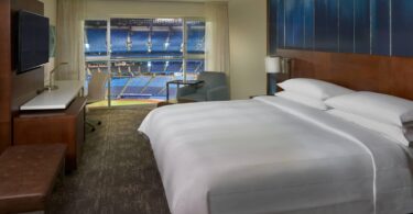 The image shows a hotel room with a large bed, a TV mounted on the wall, a desk with a chair, and a small armchair. The room has a large window with a view of a sports stadium, showing rows of blue seats and part of the field.
