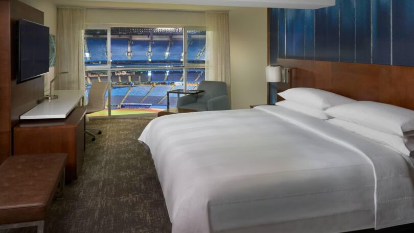 The image shows a hotel room with a large bed, a TV mounted on the wall, a desk with a chair, and a small armchair. The room has a large window with a view of a sports stadium, showing rows of blue seats and part of the field.