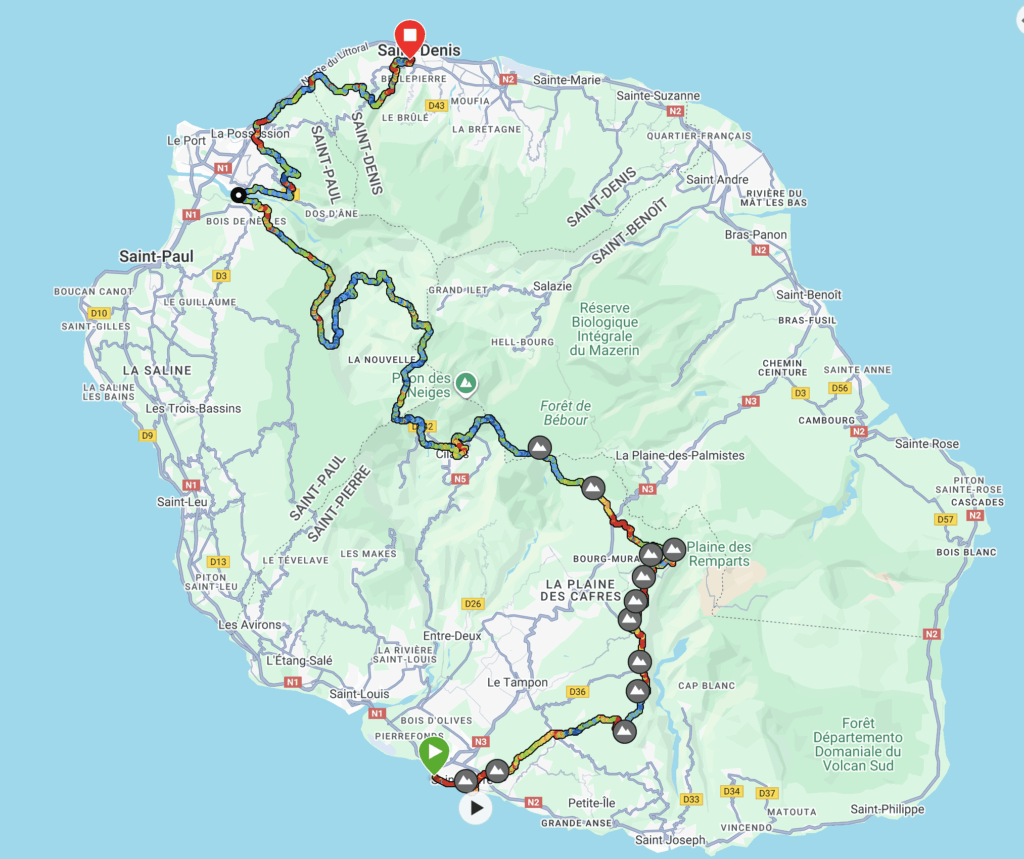 The image is a map of Réunion Island, showing a detailed route for an event or journey. The route is marked with a multicolored line that traverses various regions, including Saint-Denis, Saint-Paul, and Saint-Pierre. The map highlights roads, towns, and natural features like forests and mountains. Key points along the route are marked with icons, possibly indicating checkpoints or significant locations. The map is set against a light blue background representing the ocean surrounding the island.