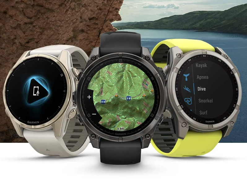 The image shows three Garmin smartwatches with different displays. The left watch has a charging icon on its screen, the middle watch displays a topographic map, and the right watch shows a menu with options like "Kayak," "Apnea," "Dive," "Snorkel," and "Surf." The watches have different band colors: white, black, and yellow. The background features a rocky surface and a scenic view of a lake with hills.