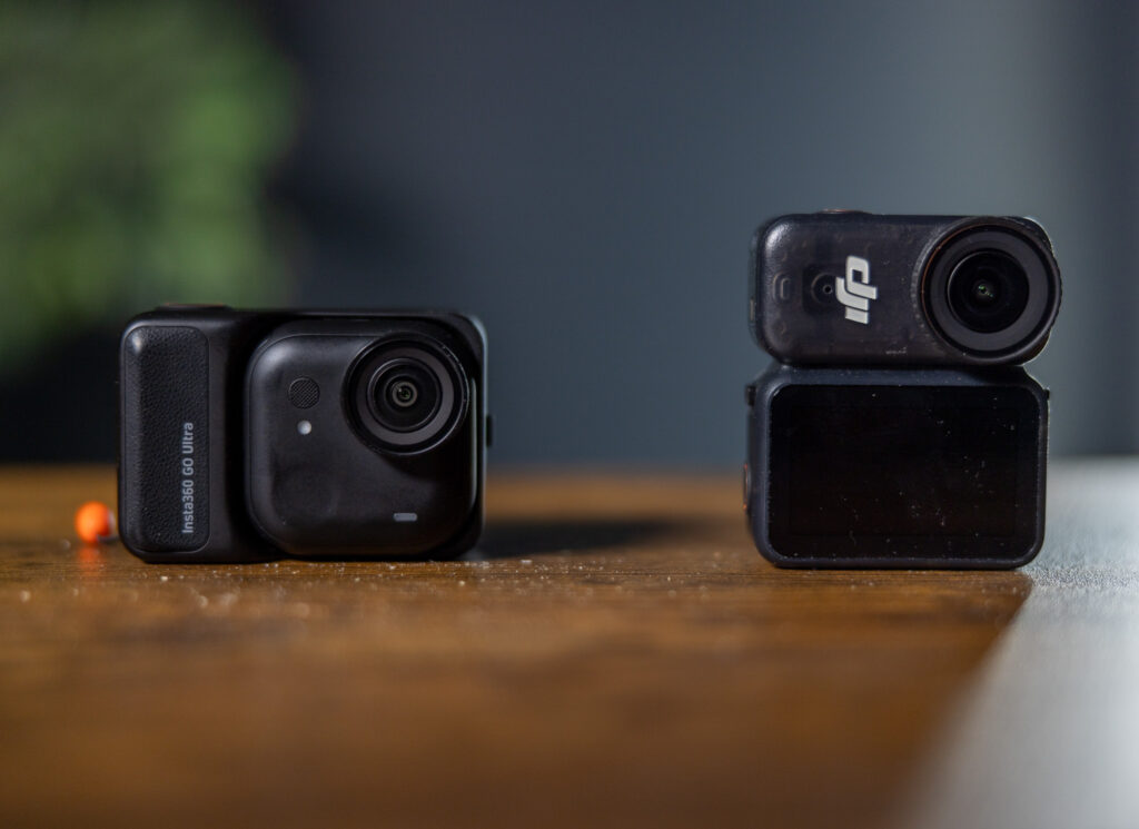 The image shows two compact action cameras placed on a wooden surface. The camera on the left is labeled "Insta360 GO Ultra" and has a small orange object next to it. The camera on the right has a lens on top and a screen on the bottom. The background is blurred, with a hint of greenery on the left.