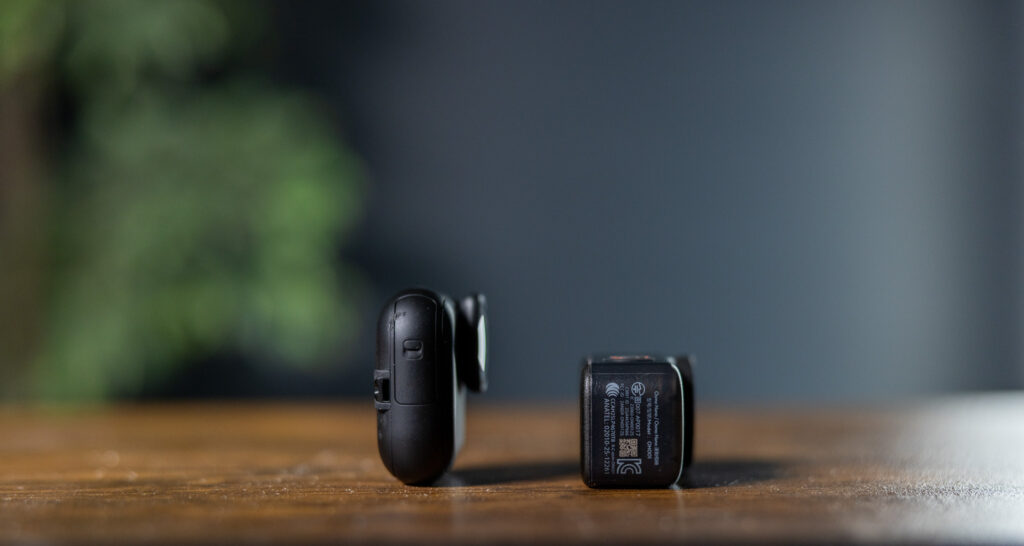 The image shows two small black electronic devices placed on a wooden surface. One device is standing upright, while the other is lying on its side. The background is blurred, with hints of green and gray tones.