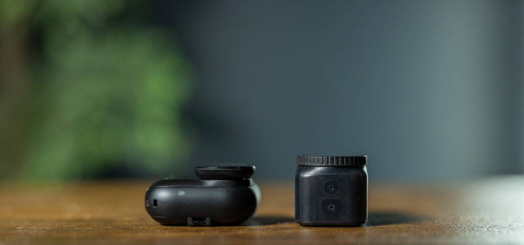 A small black camera lens and a black lens cap are placed on a wooden surface. The background is blurred with green and gray tones.
