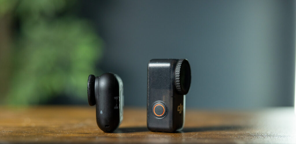 The image shows two small black camera devices placed on a wooden surface. The device on the left is compact with a rounded design, while the device on the right is slightly larger with a lens and a button featuring an orange ring. The background is blurred with green and gray tones.