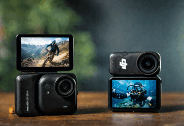 The image shows two action cameras placed on a wooden surface. The camera on the left has a screen displaying a person mountain biking on a rocky trail with mountains in the background. The camera on the right shows a screen with a person scuba diving underwater. Both cameras have lenses prominently visible, and the background is blurred.