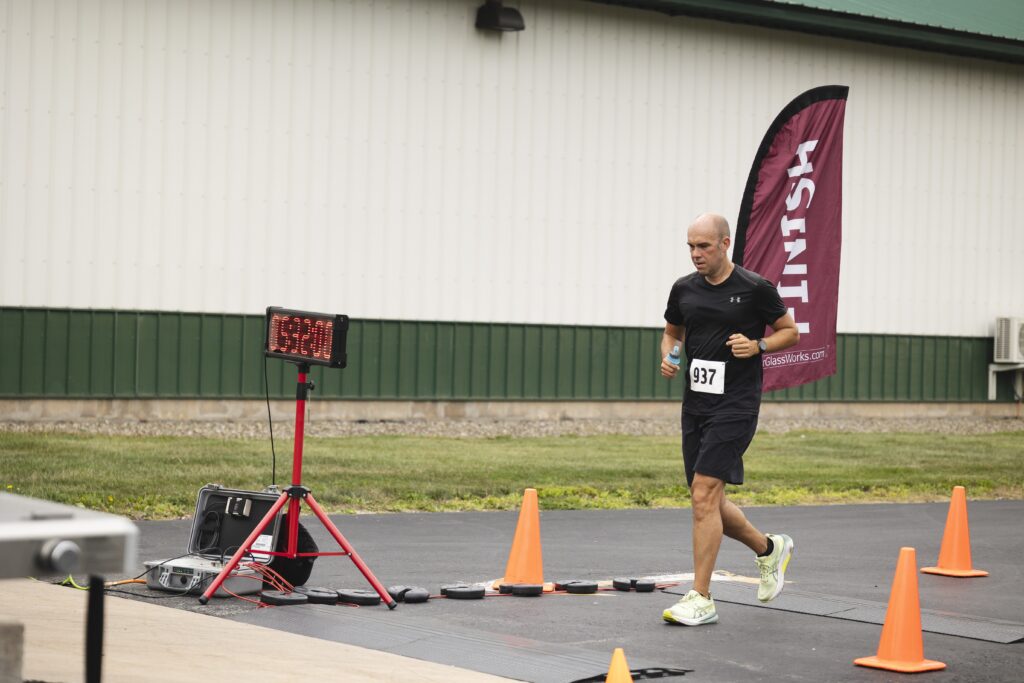 A man is running towards a finish line during a race. He is wearing a black shirt, black shorts, and running shoes, with a race bib number 937. He is holding a small water bottle. There is a digital timer on a stand showing "03:30:00" and a maroon flag with the word "FINISH" written on it. Orange cones are placed on the ground, and a building with a green and white exterior is in the background.