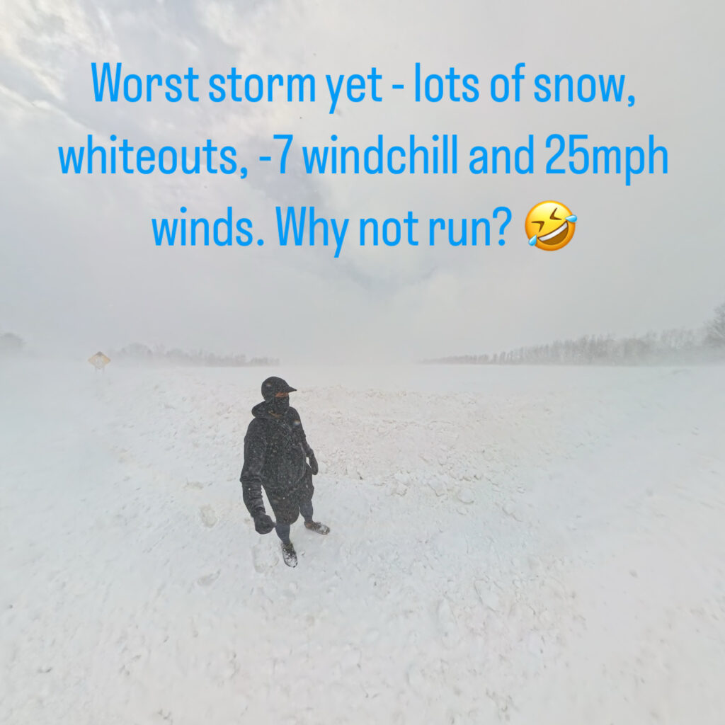 A person is standing in a snowy landscape during a severe winter storm. The ground is covered in snow, and visibility is low due to whiteout conditions. The person is dressed in winter clothing, including a hat and gloves. The sky is overcast. Text on the image reads: "Worst storm yet - lots of snow, whiteouts, -7 windchill and 25mph winds. Why not run?" followed by a laughing emoji.